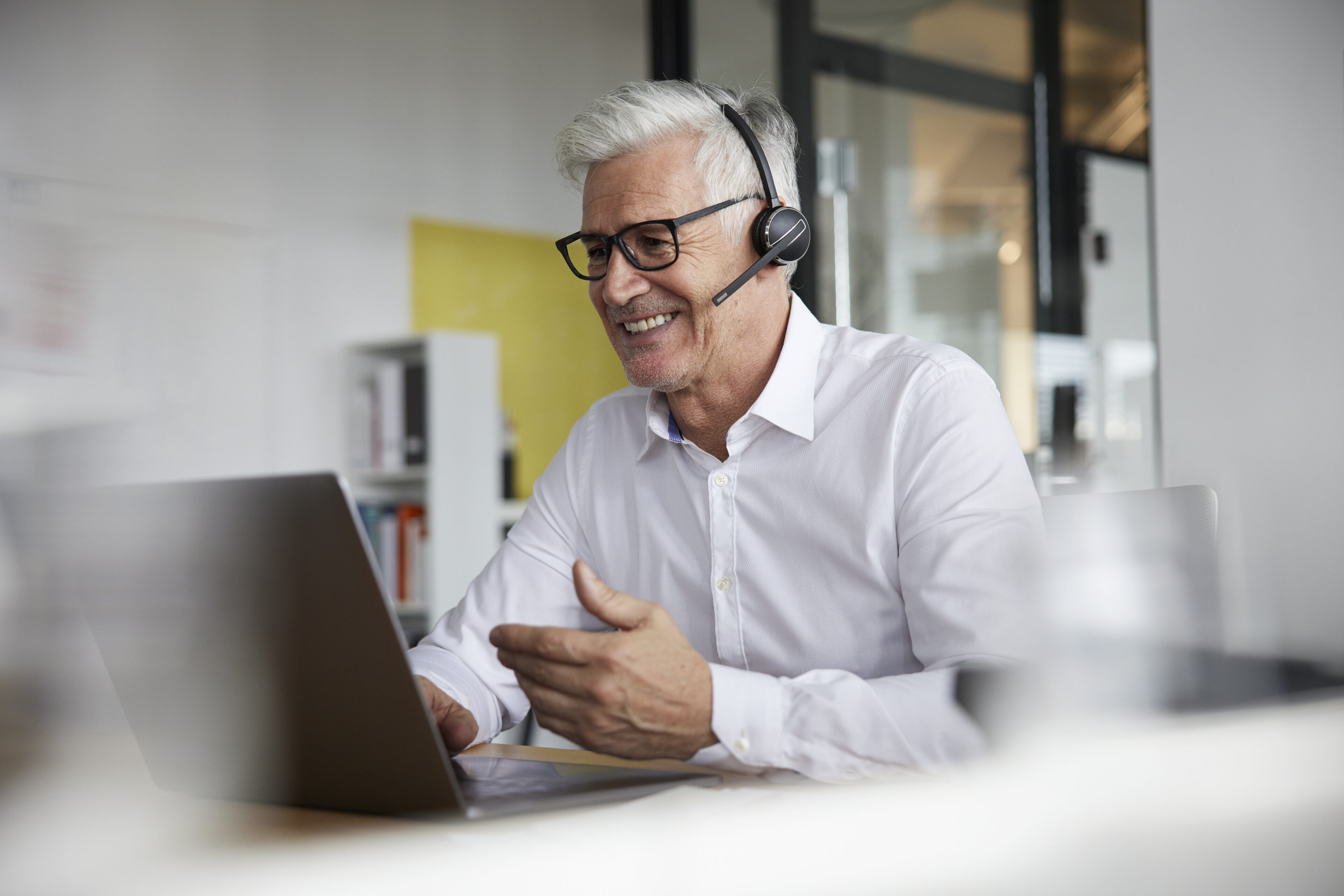 Smiling businessman with headset gesturing while talking to video call on laptop in office